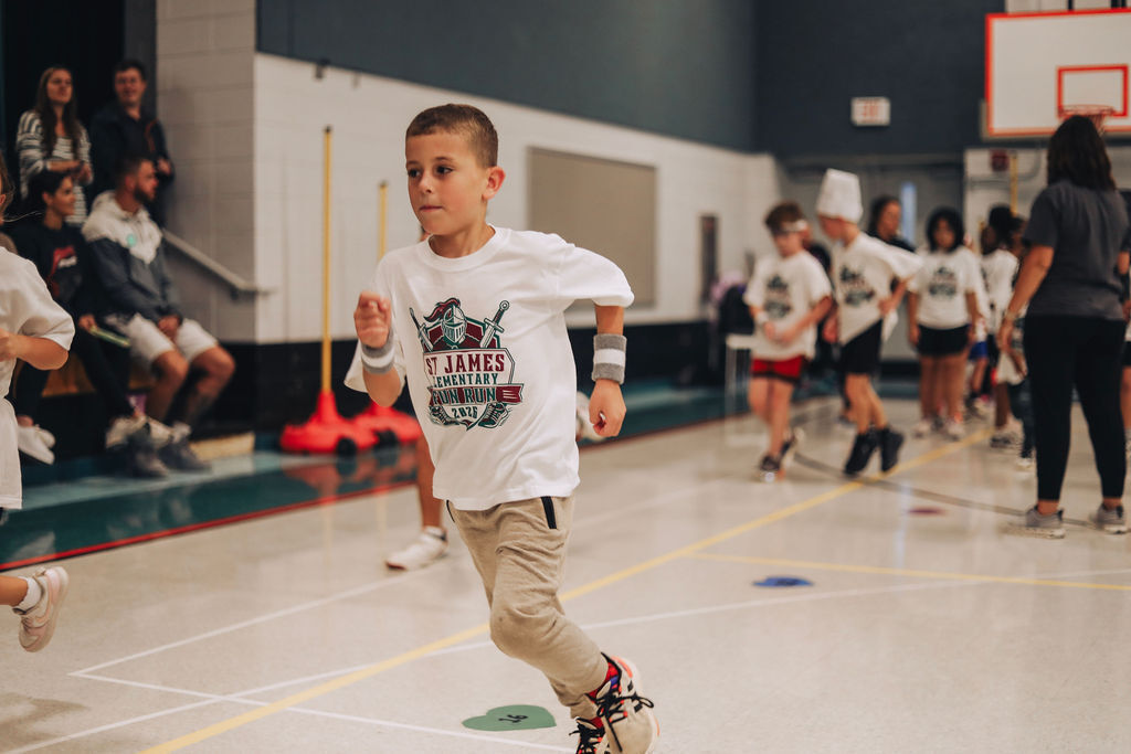 A young boy runs on a gym floor, wearing a white shirt with a graphic design. Other children in white shirts and shorts follow behind.