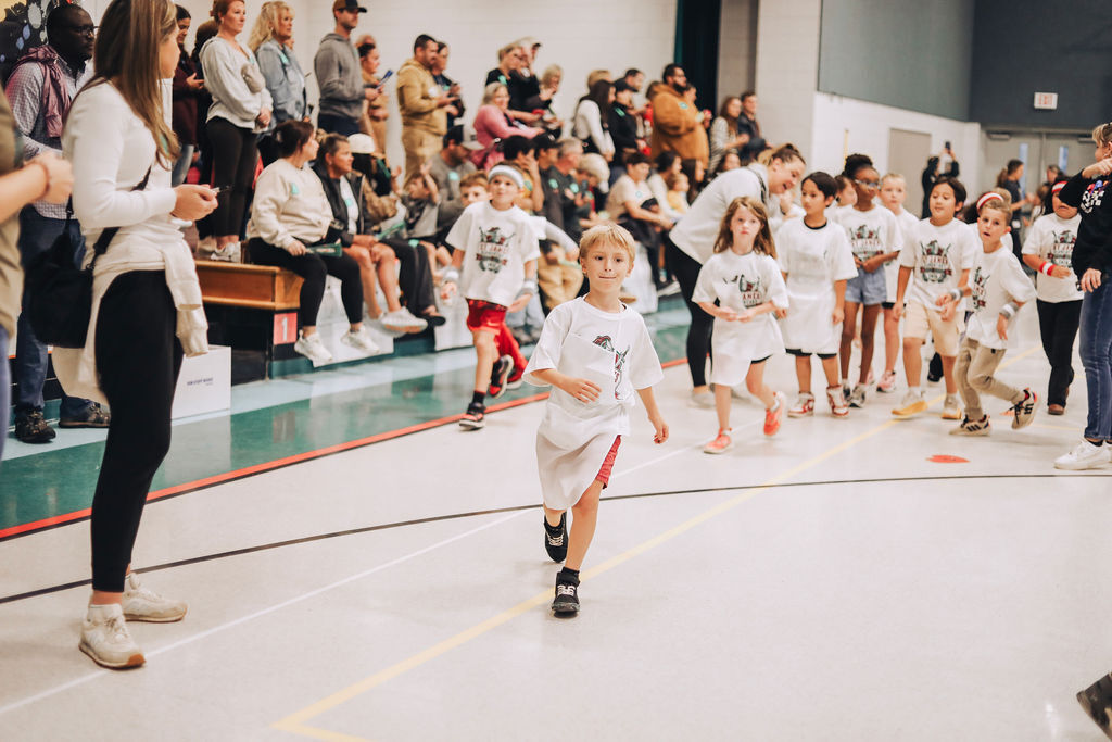 Children in white shirts and shorts running on a track with spectators seated on bleachers.