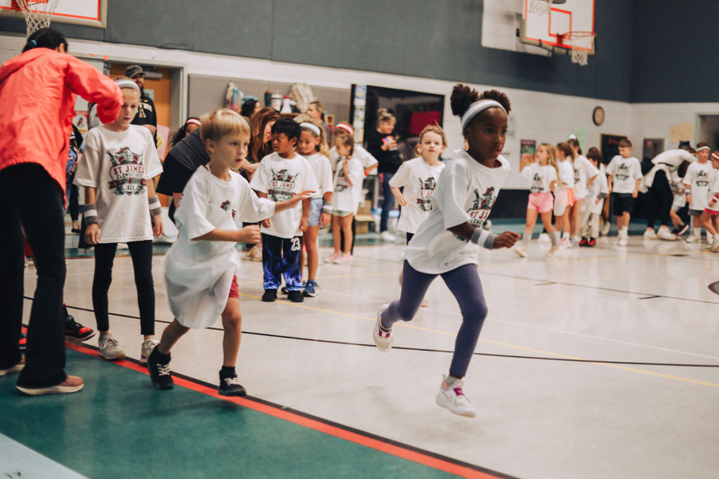 Children in white shirts run in an indoor basketball court. A person in orange stands behind them.
