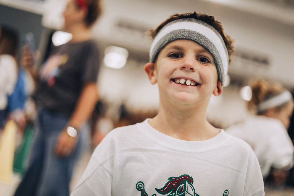 A young boy with a white headband and a white shirt with green graphics smiles at the camera. Blurry people are in the background.