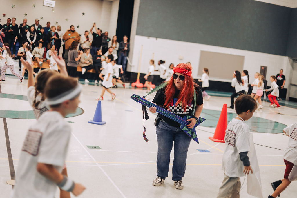 A woman with red hair and a red bandana leads a group of children in a gym.