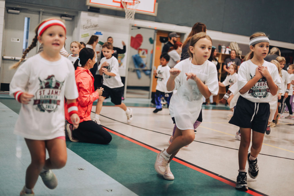 Several children in gym clothes run in a gymnasium; a woman crouches nearby. A basketball hoop and colorful murals are in the background.