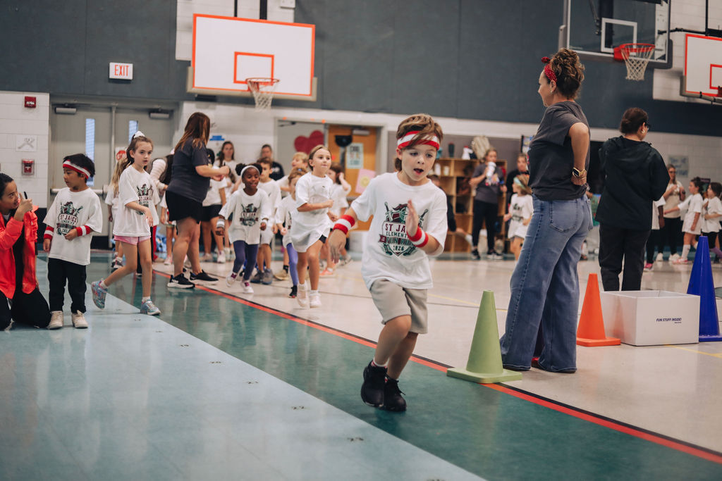 Children wearing headbands and T-shirts run along a marked path in an indoor basketball court.