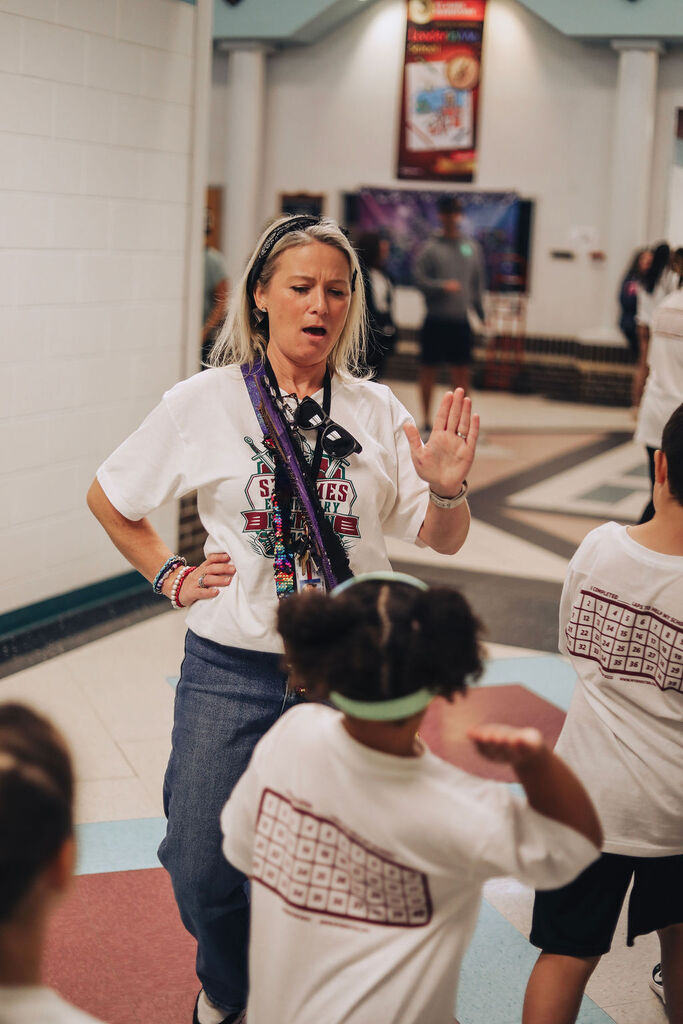 Woman with short blonde hair leads children in a hallway. She wears a white shirt with blue text and blue jeans.