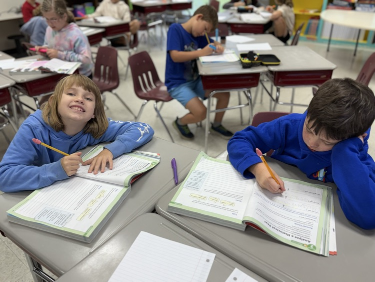 Two elementary students sit at desks side by side working in workbooks with pencils. The girl on the left wears a light blue hoodie and smiles toward the camera, while the boy on the right wears a bright blue hoodie and leans his head on his hand as he writes. Other students work at desks in the background classroom setting.