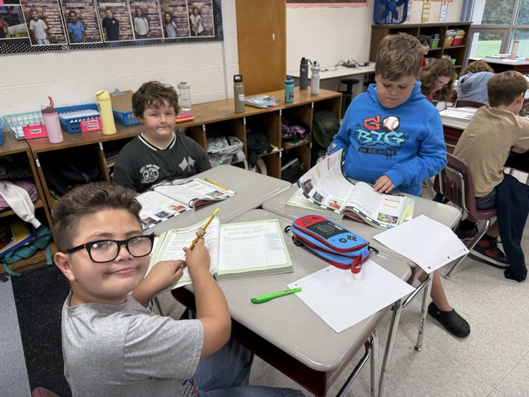 Four elementary students sit around a cluster of classroom desks engaged in schoolwork. In the foreground on the left, a boy wearing dark-rimmed glasses and a grey shirt smiles toward the camera holding a pencil over his open textbook. Beside him sits another boy in a dark t-shirt looking forward, while across the desks, a boy in a blue hoodie flips through an open workbook next to a red and blue pencil case.