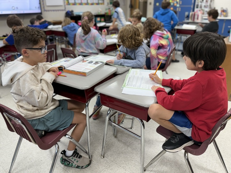 Two young students work at adjoining school desks in a classroom. On the left, a boy wearing glasses and an off-white hoodie sits facing sideways in his chair. On the right, a boy in a red long-sleeve shirt writes in a workbook with a pencil. Other students are seated at desks working in the background classroom setting.