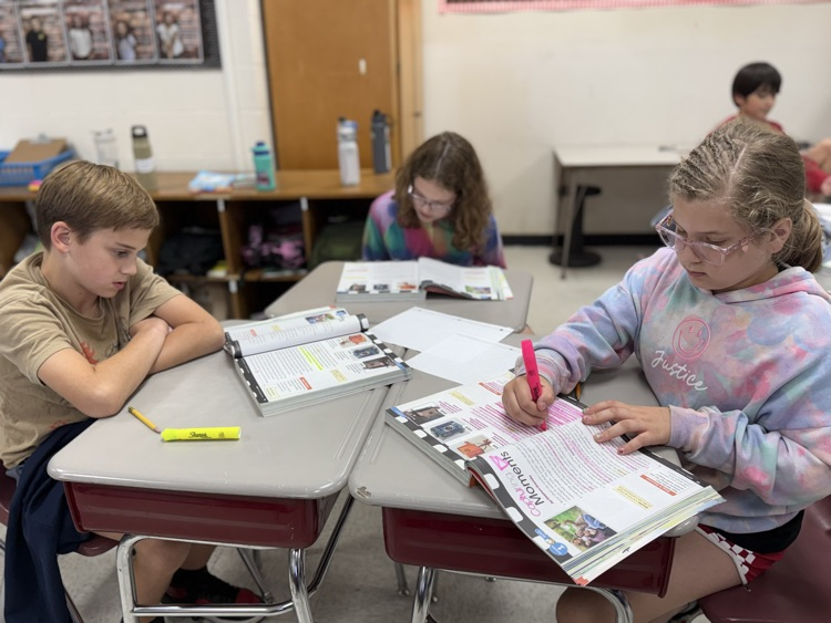 Two elementary students sit side by side at classroom desks reading and writing in open textbooks. The girl on the right wearing clear-framed glasses and a pastel tie-dye hoodie writes in her book with a pink marker, while the boy on the left in a tan shirt sits with arms crossed looking down at his desk. In the background, another student with dark hair sits at a desk studying.