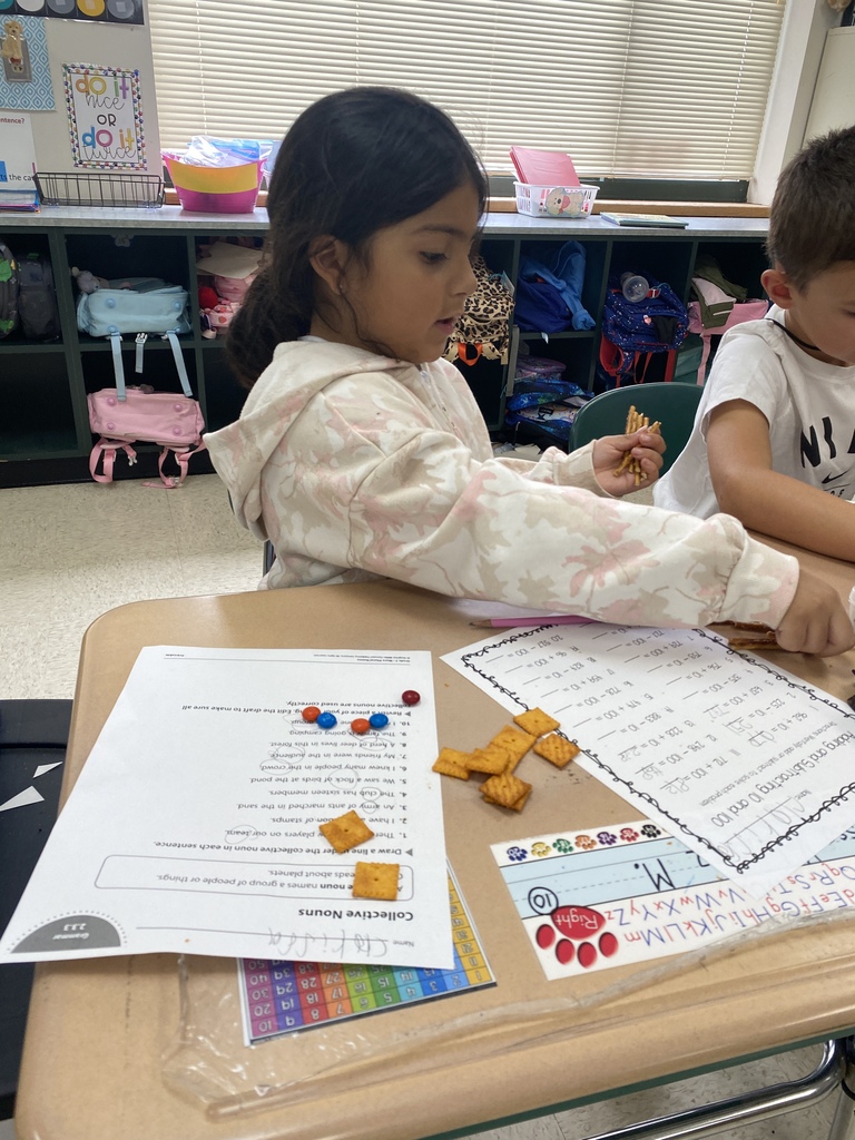 Two children seated at a desk, one girl is reading a paper with numbers, and both have small objects on the table.