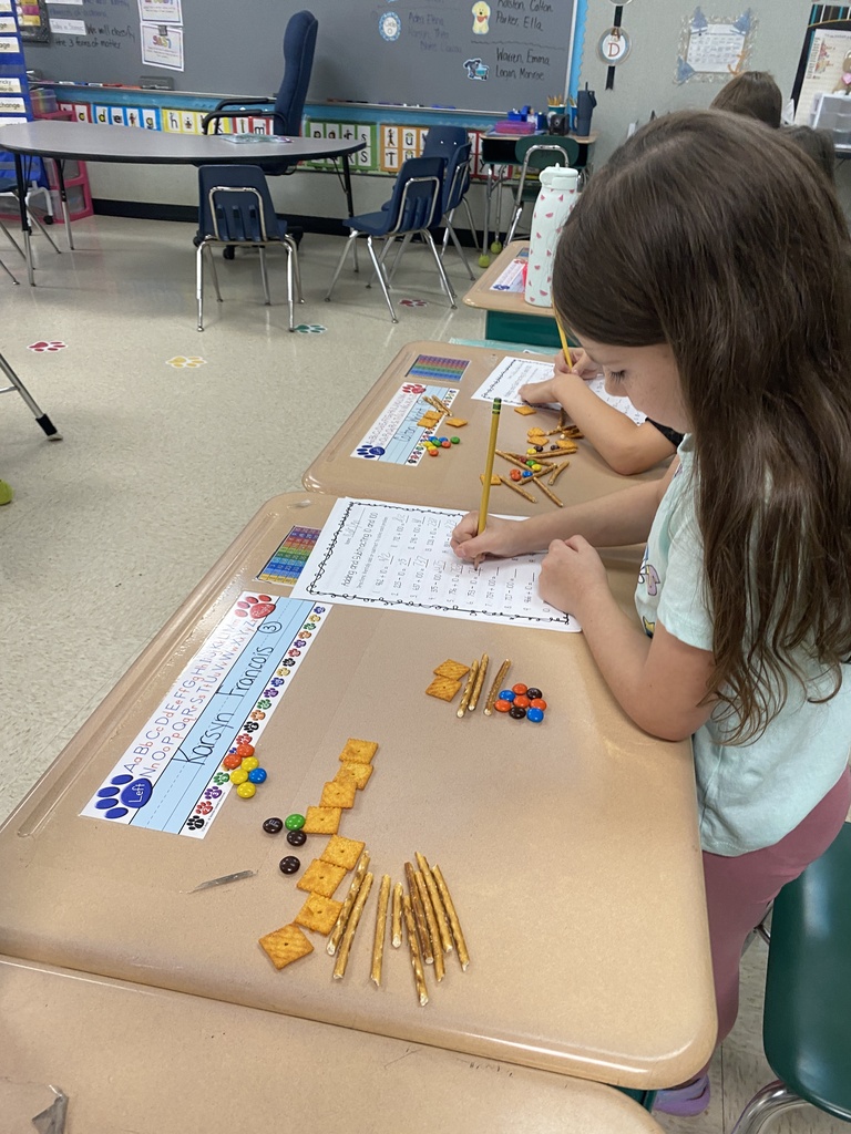 A girl at a desk in a classroom uses blocks to help her with math. Behind her, another student works at a separate desk.