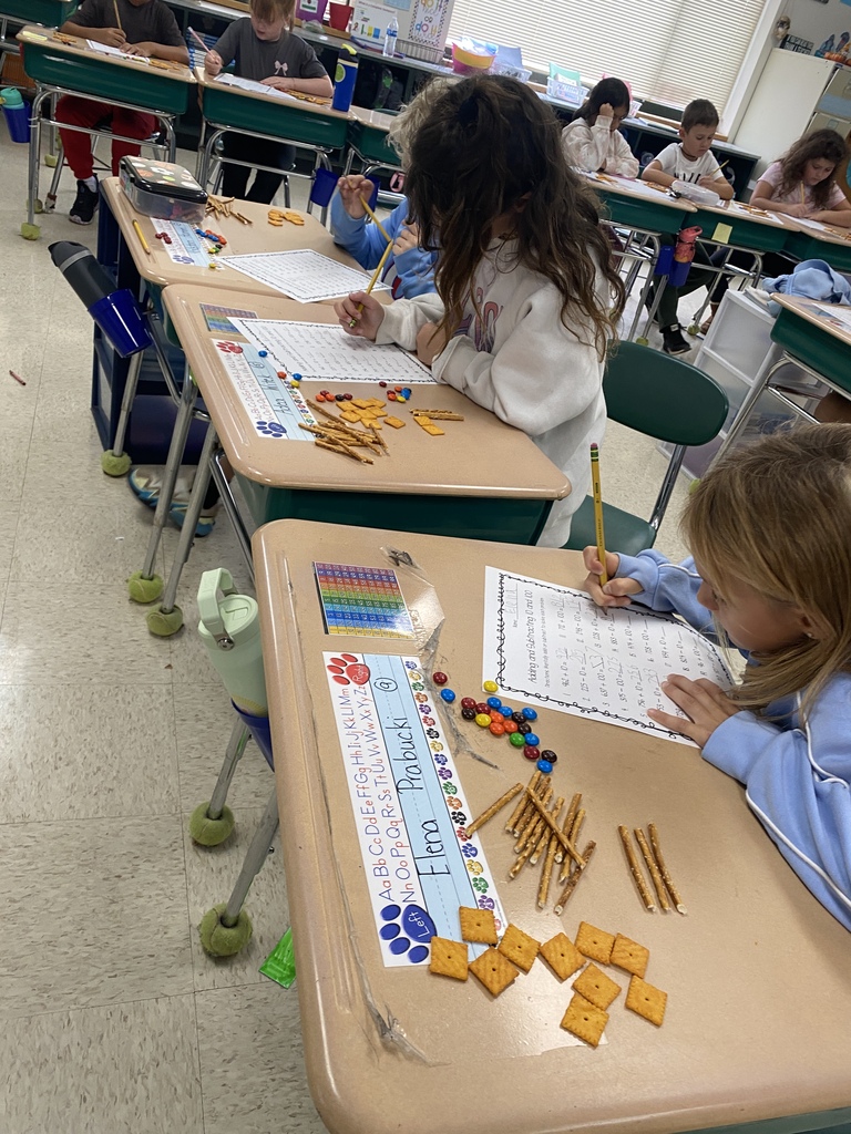 Children seated at desks in a classroom, using educational materials like pencils and blocks. One child has a snack, and the desks are equipped with colorful papers.