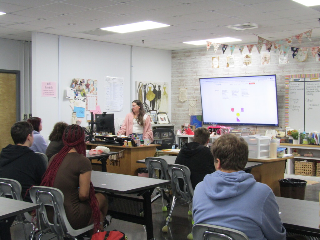 A classroom with a woman standing at a podium. People seated at tables face her. The room has whiteboards and a large screen.
