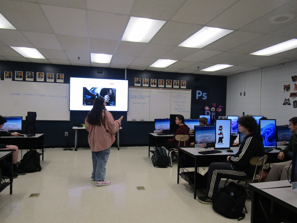 A classroom with students seated at desks, a person standing near the front with a projector screen behind them.