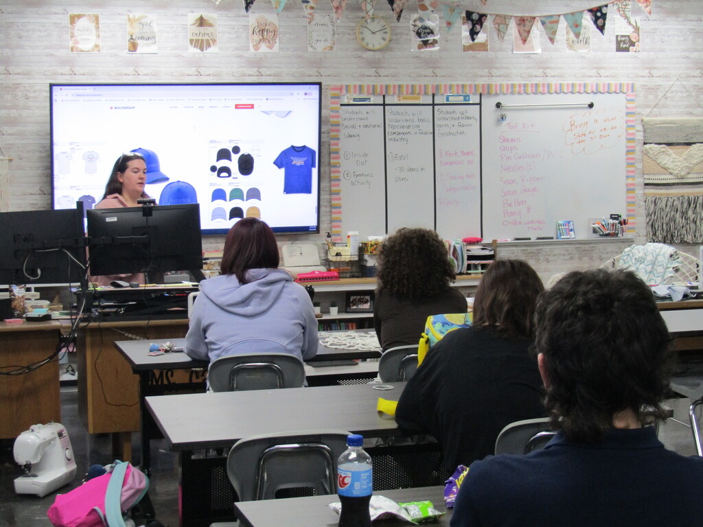 A classroom with a teacher at a computer screen. Students sit at desks, facing the screen. Whiteboards and various classroom items are visible.