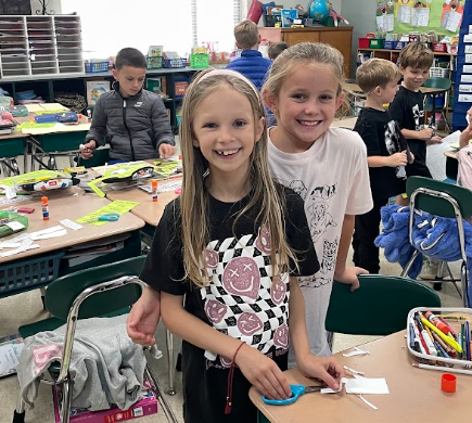 Two girls, one wearing a black t-shirt, smile in a classroom. Desks, chairs, and students are in the background.
