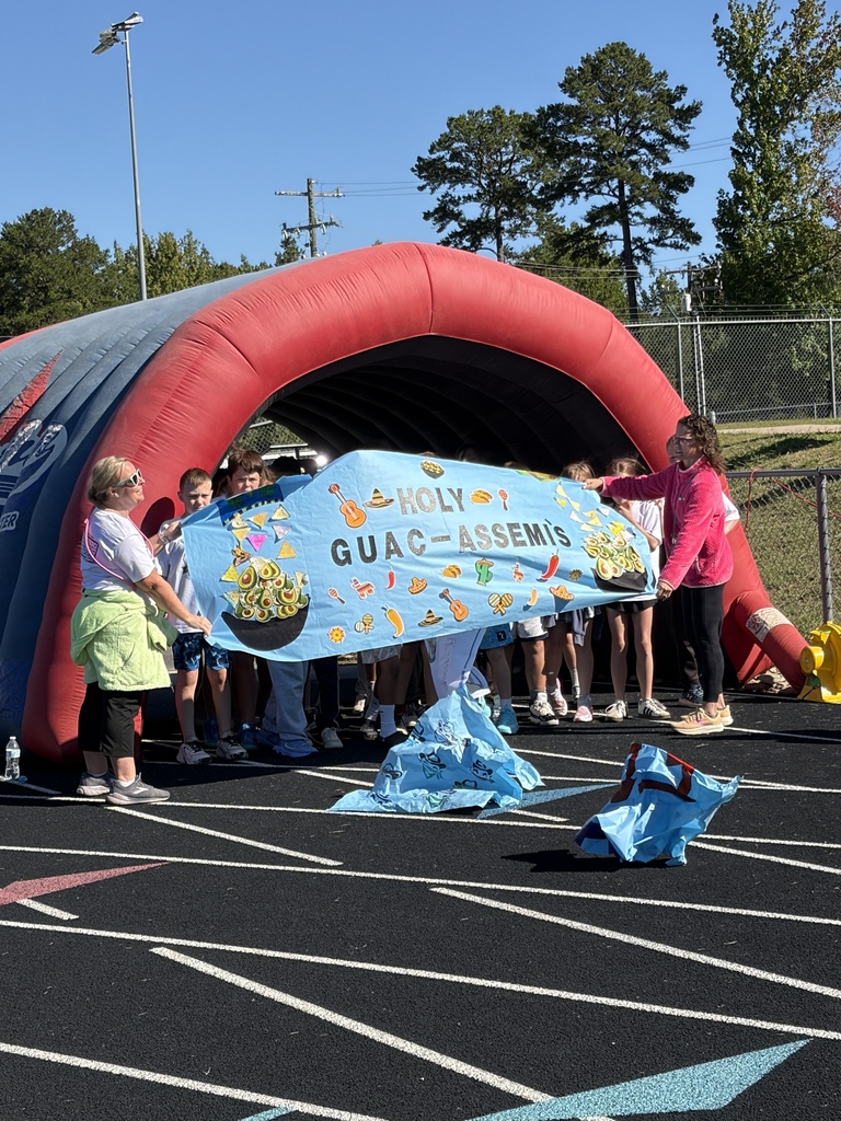 A group of children holds a banner for Holy Guacamole Assembly under a large arch on a field.