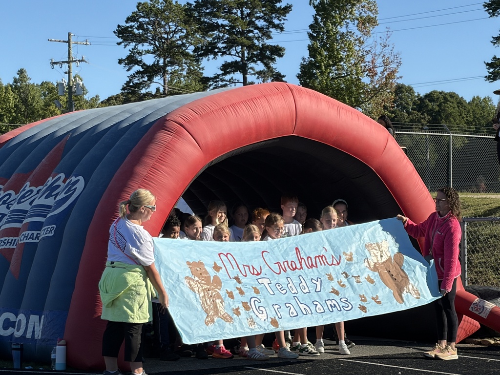 People gather under a large inflatable tent, with two adults holding a banner. The banner reads "Mr. Graham's Team". Trees and a fence are in the background.