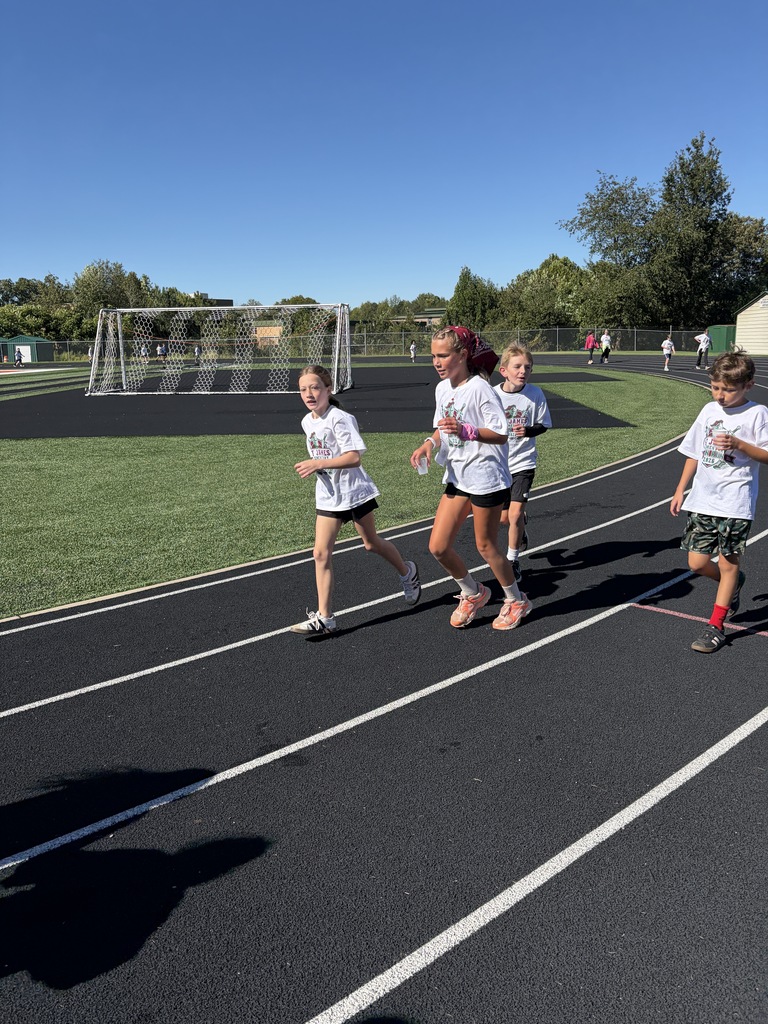 A group of children running on a track. They are wearing white shirts. A grassy field and a soccer goal are in the background.