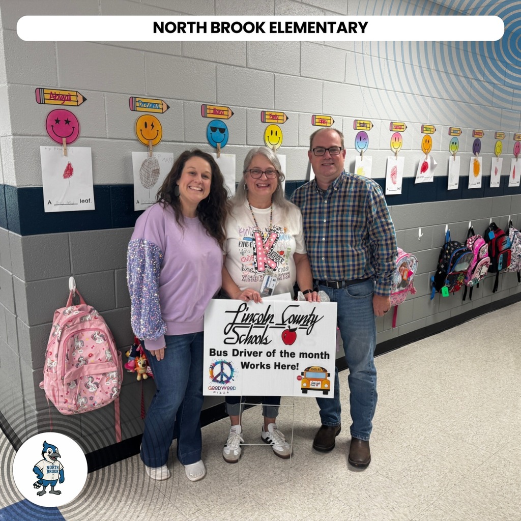 Three people stand in a hallway. One holds a sign. A backpack and school supplies hang in the background.