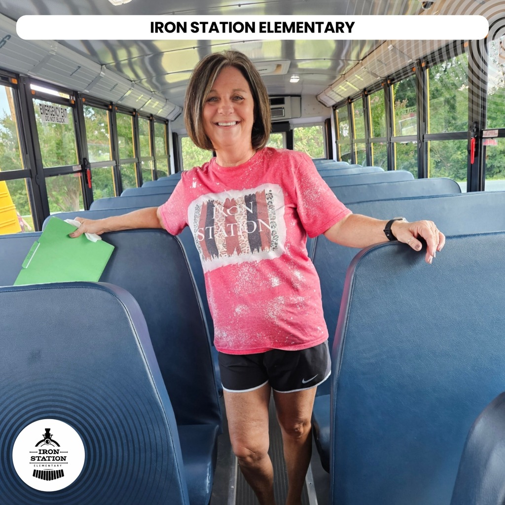 A woman wearing a pink shirt and black shorts smiles while standing on a school bus.