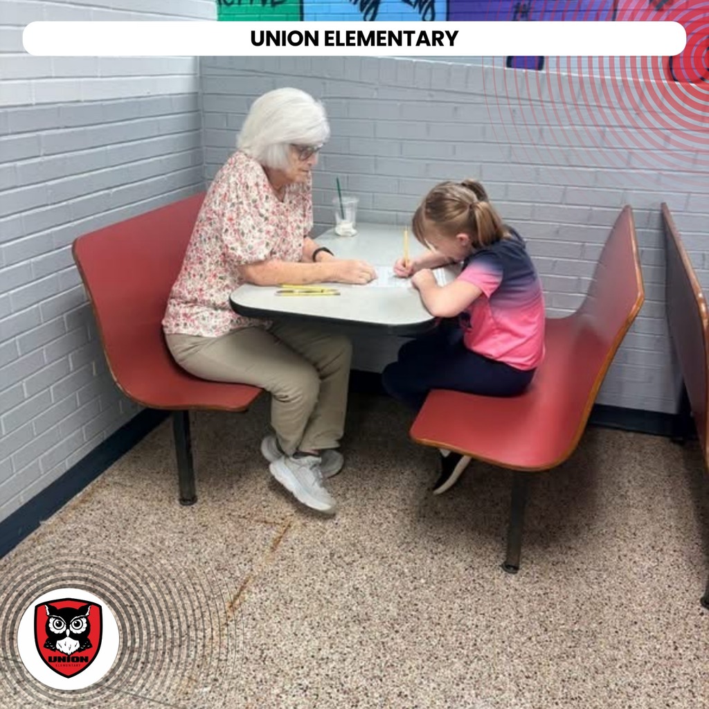 An elderly woman and a child are seated at a table. The woman is wearing a floral shirt and the child a pink shirt.