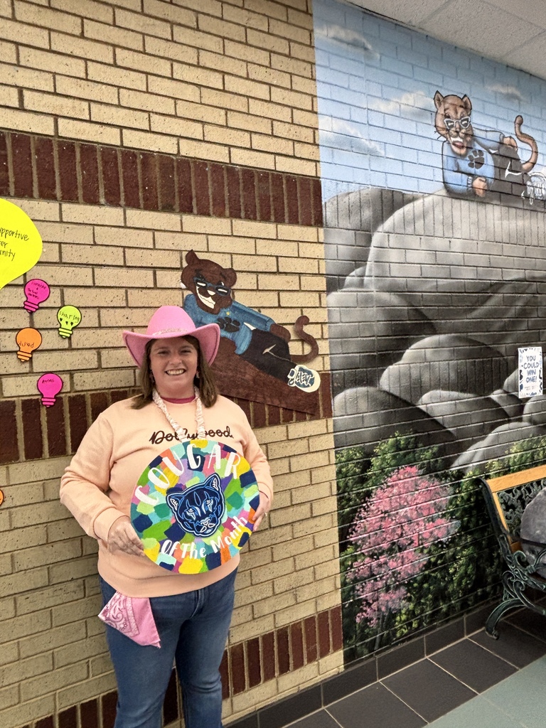 Woman standing in a school hallway wearing a pink cowboy hat, peach sweatshirt, and jeans while holding a colorful round sign that reads "Cougar Of The Month" with a cougar mascot illustration. She stands next to a brick wall with lightbulb cutout signs and a painted wall mural featuring a cougar mascot perched on a rock above trees.