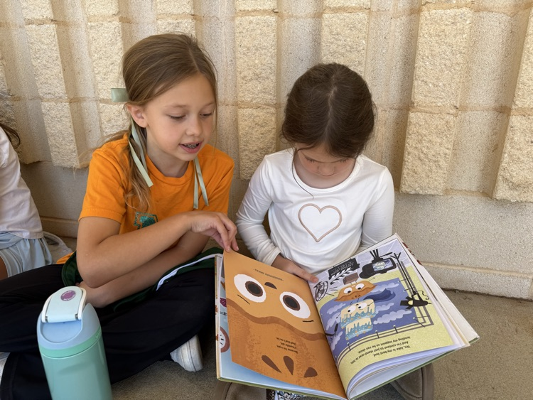 Two young girls sit side by side on the floor against a stone wall reading an open illustrated picture book together. The girl on the left wears an orange shirt and turns a page while pointing at an illustration, while the girl on the right wears a long-sleeve white shirt with an outlined heart on the front and looks down at the book.