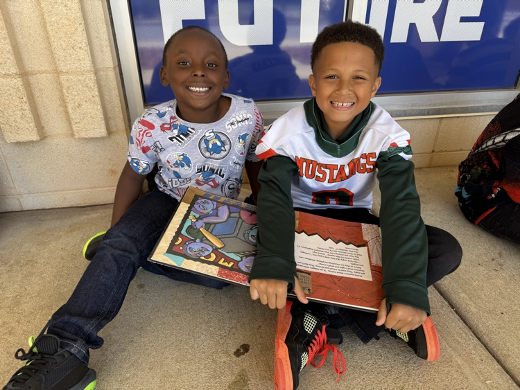 Two smiling young boys sit side-by-side on the concrete ground outside against a wall beneath a blue sign. The boy on the left wears a patterned grey Sonic the Hedgehog shirt and dark pants, while the boy on the right wears a white and green Mustangs jersey over a long-sleeve shirt. Together, they hold an open picture book across their laps.