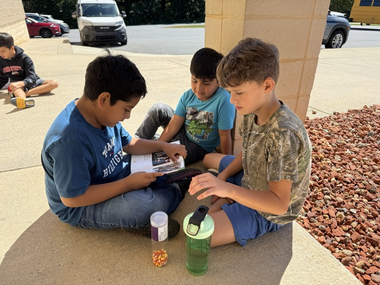 Three young boys sit together outdoors on the sidewalk against a stone column reading a book. The boy on the left in a blue Tommy Hilfiger shirt holds open a small paperback book, while a boy in a teal shirt and a boy in a camouflage shirt lean in to look at the pages. A clear container of candy corn and a green water bottle rest on the ground in front of them.