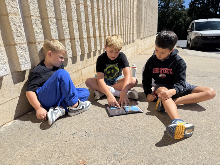 Three young boys sit together cross-legged outdoors on the concrete ground against a stone wall reading a open book placed on the ground. The boy in the middle points to the text in the book while the boys on either side look on. The boy on the left wears a black shirt and blue pants, the middle boy wears a black graphics shirt and shorts, and the boy on the right wears a dark Ohio State hoodie and shorts.
