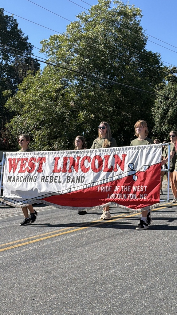 People wearing sunglasses, holding a banner reading "West Lincoln Marching Rebel Band," walk on a road. Trees and power lines in the background.