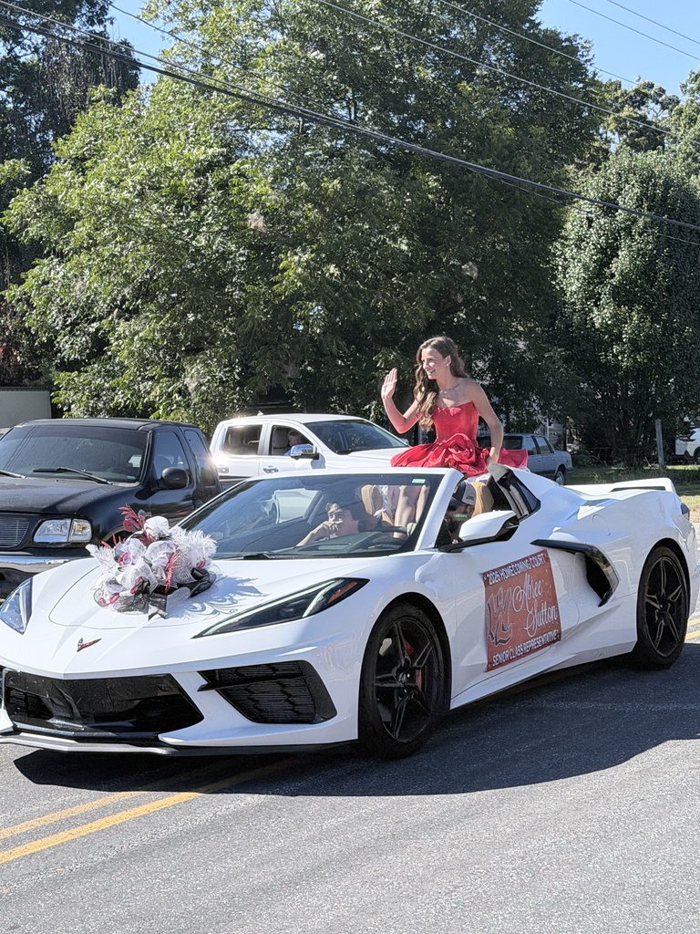 A white sports car with a woman in a red dress drives on the road with trees and other vehicles in the background.