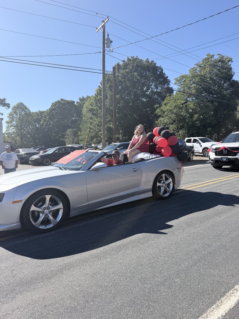 Convertible car with several people riding it, two with balloons, driving on road with trees in background.