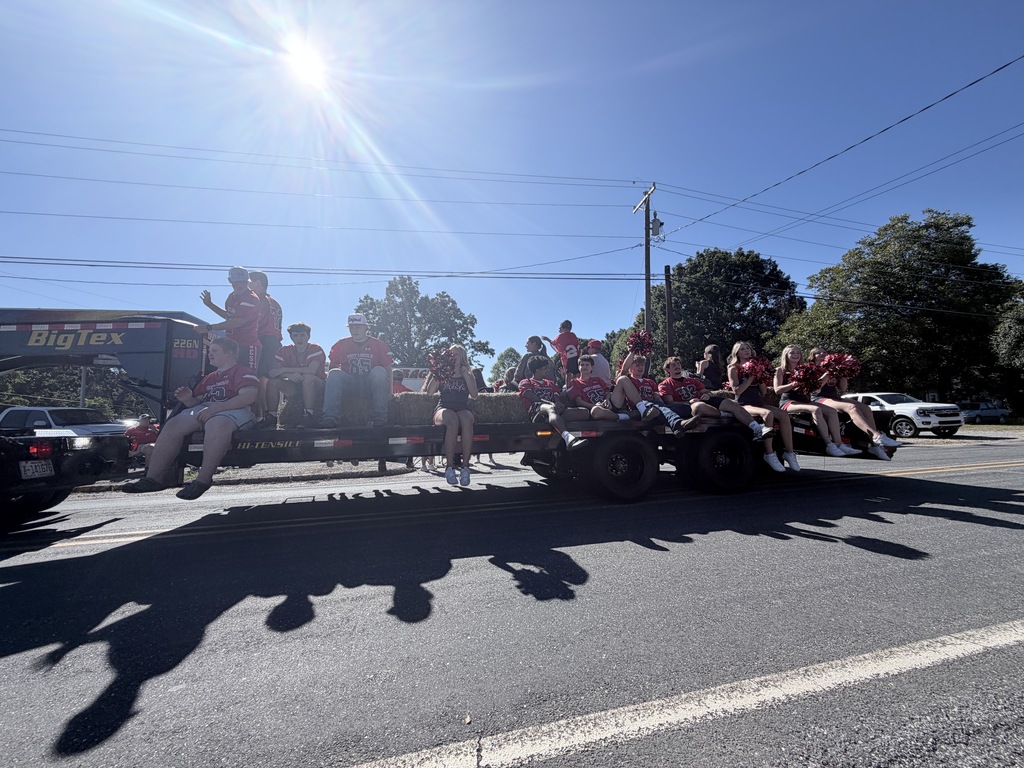 A group of people in red attire on a trailer during a parade. Trees and power lines in the background.