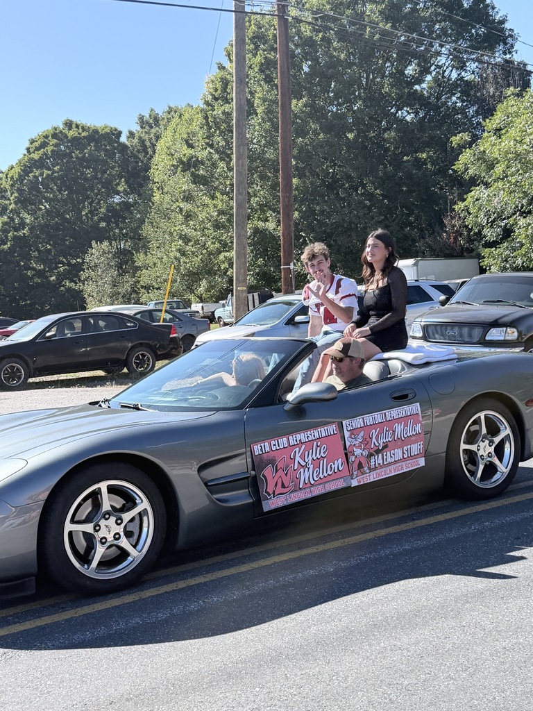 A gray convertible car in a parade with three people in it, one wearing a hat, passes by parked cars and trees.