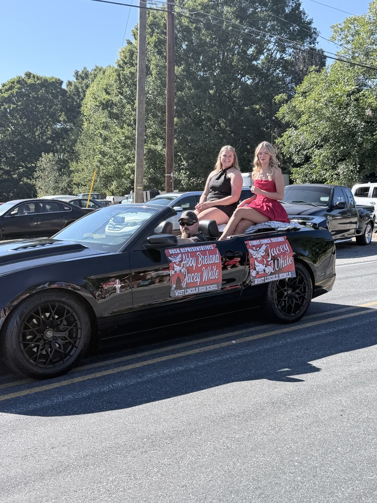 A convertible vehicle with two women in red dresses on the back seats. A road is visible with trees and parked cars.
