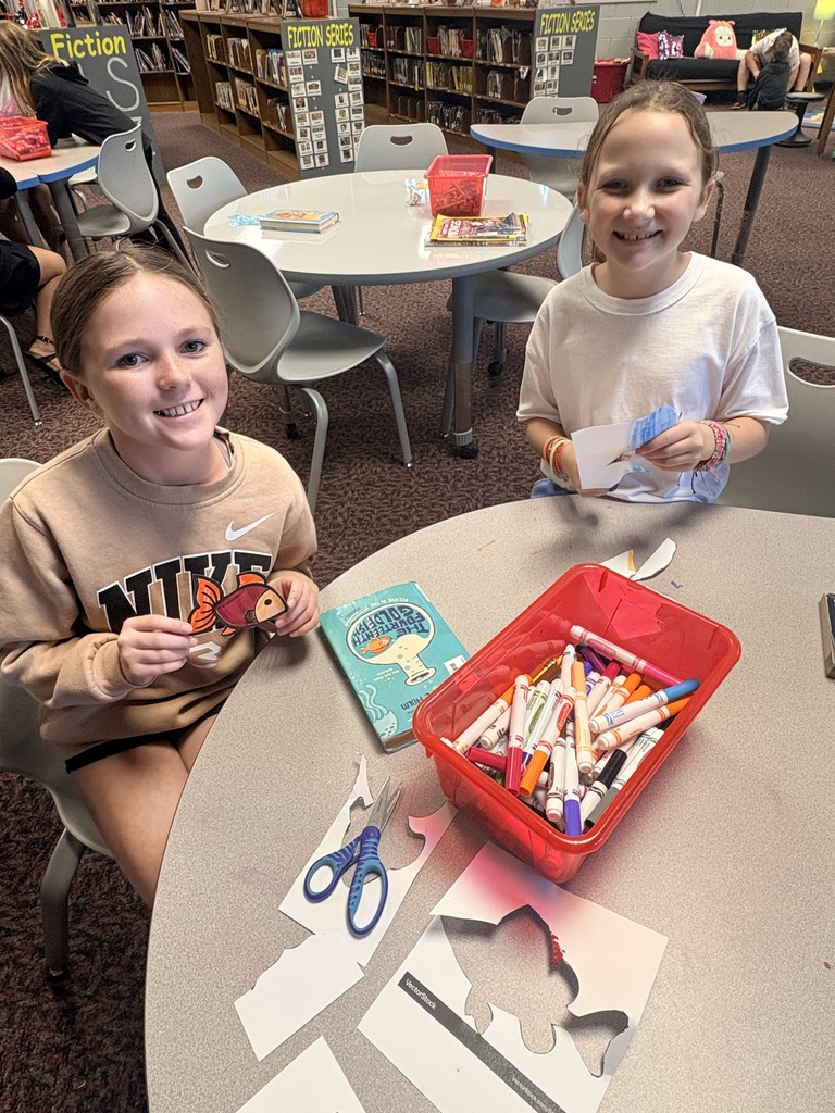 Two girls seated at a table. One holds scissors, another holds paper. A red bin of markers and a book nearby. Background: library.