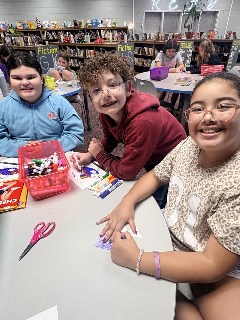 Three students in a library setting, seated at a table. One holds scissors, one a box of markers, and all are smiling.