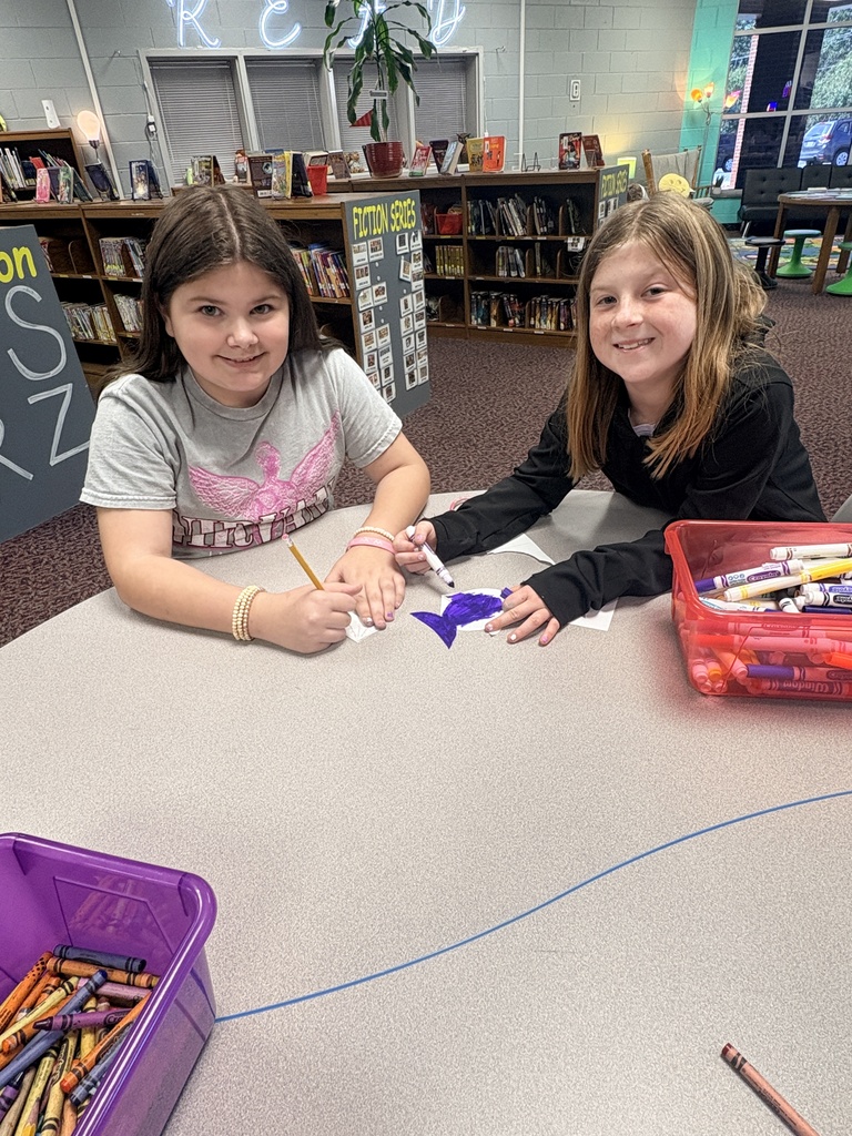 Two children at a table, one holding a crayon and the other a pencil, coloring. Behind them are shelves with books.