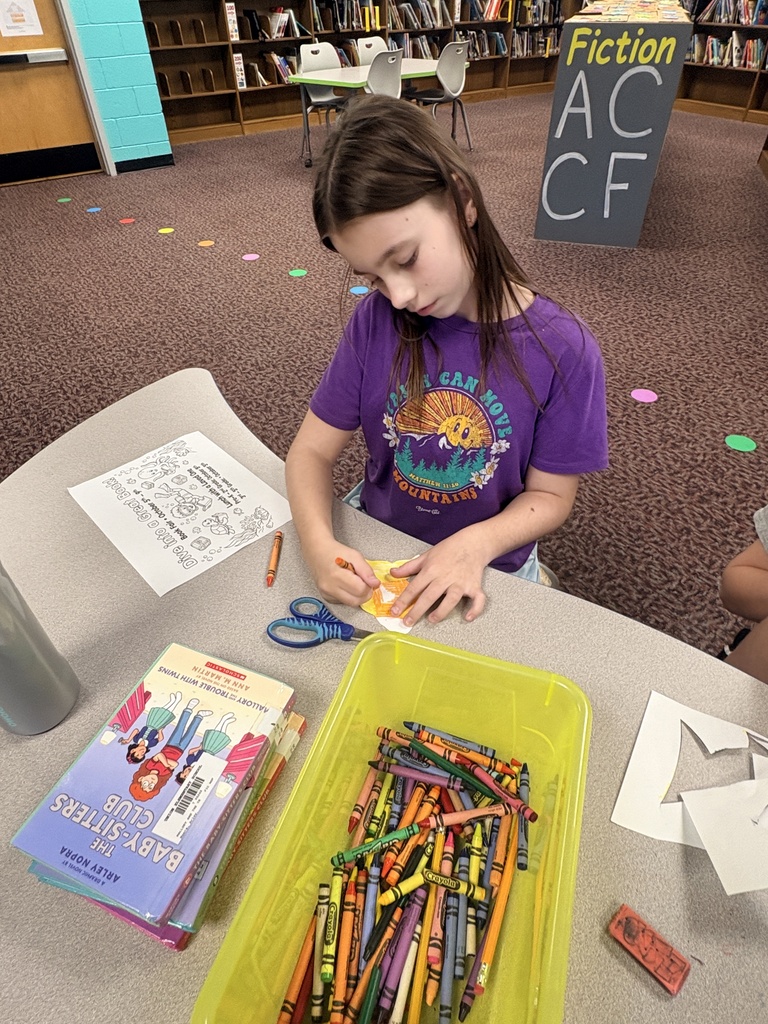 A girl in a purple shirt sits at a table in a library, working on a craft with crayons.