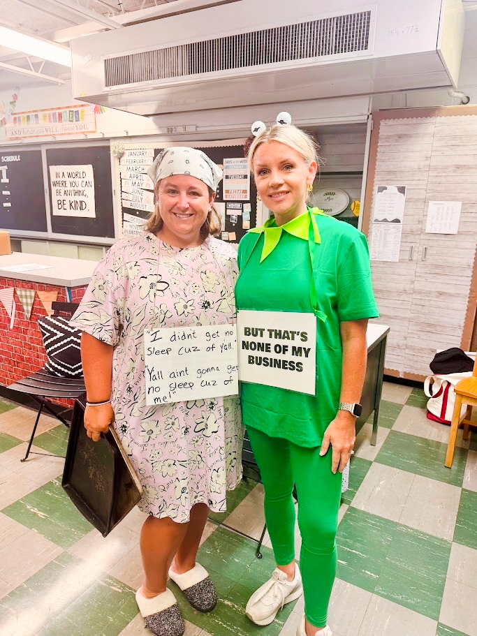 Two adults dressed in Halloween costumes, one in green scrubs, the other in a white dress. They stand in a classroom.