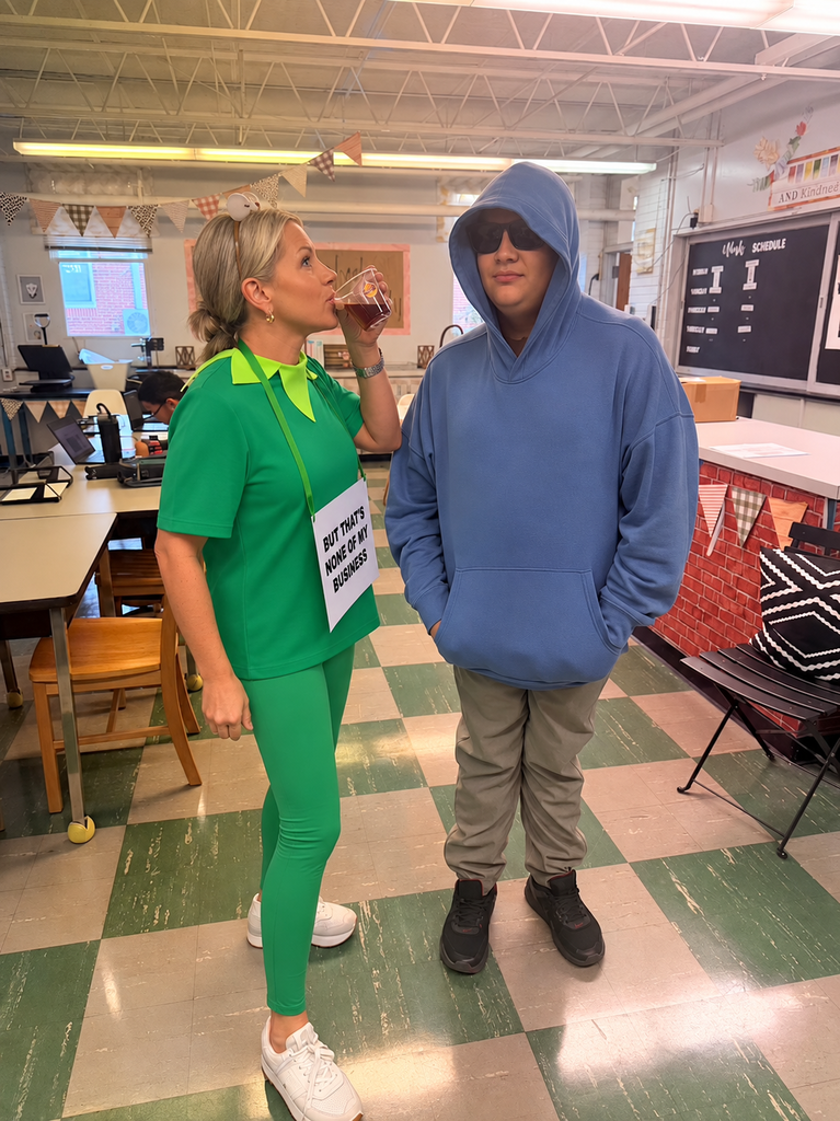 A woman in green with a sign and a man in blue are standing in a room. The floor is checkered.