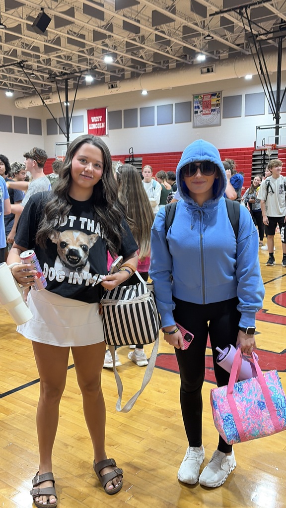 Two women in a gym, one wearing a black shirt with dog design, white skirt, and sandals, holding a water bottle. The other in a blue hoodie, black pants, and sneakers, carrying a pink bag.