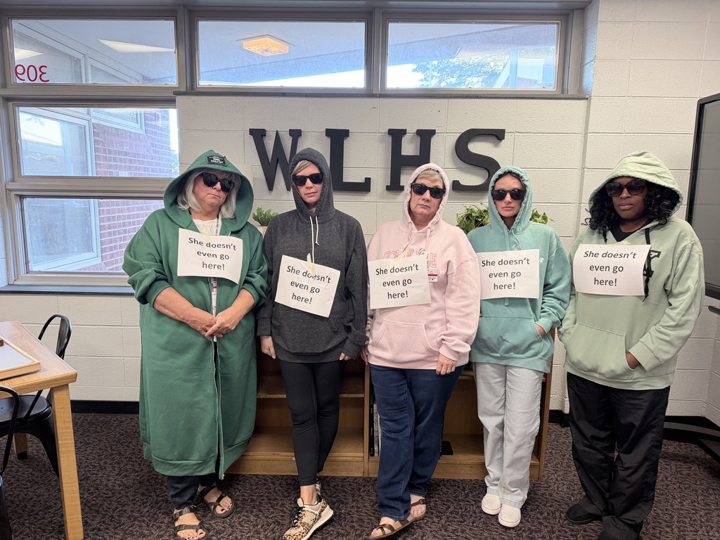 Five people in matching hoodies stand together in front of a sign that reads WLHS. They hold signs and pose for a photo.