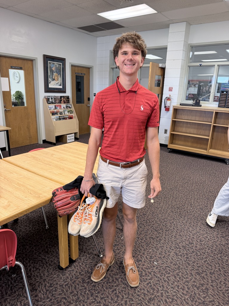 Man in red polo shirt and shorts holding shoes and smiling. Behind him is a wooden table, a red chair, and a fire extinguisher.