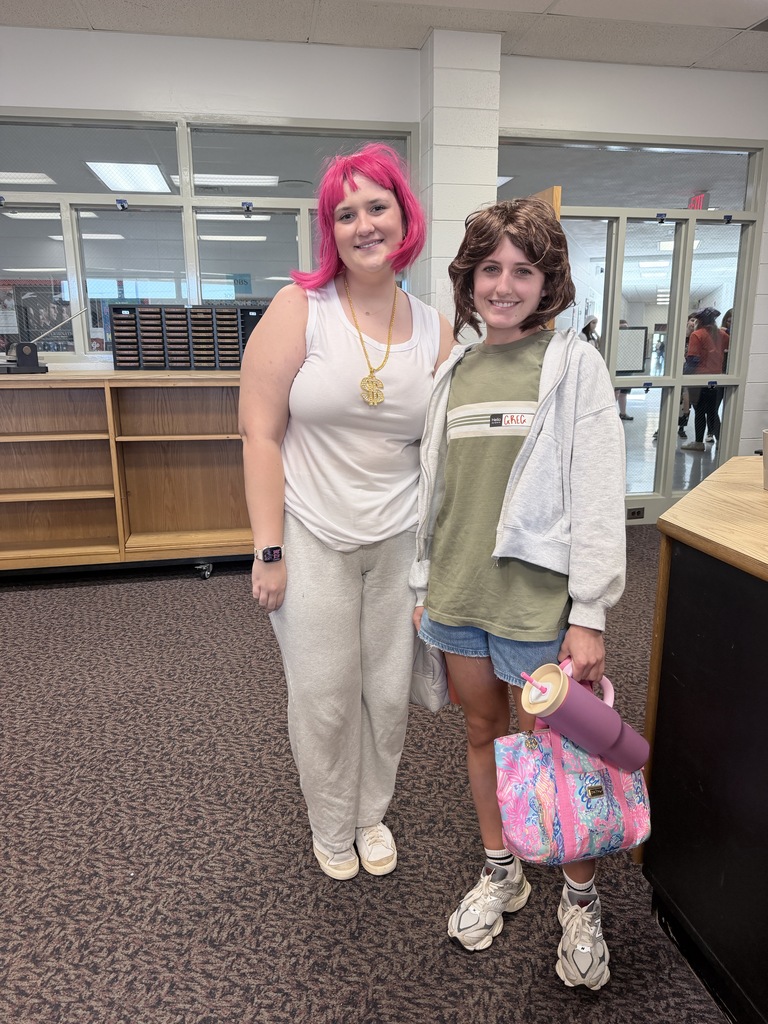 Two individuals in a library. One wears pink hair and white pants; the other wears a green shirt, shorts, and a pink bag.