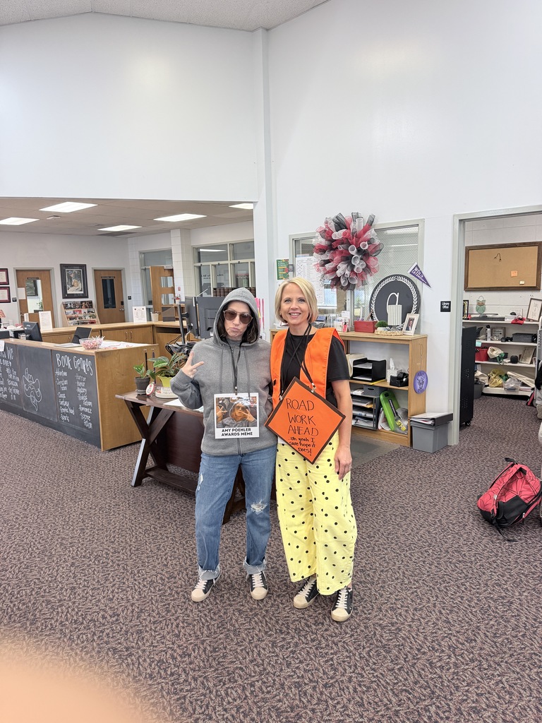 Two people in costumes stand in an open room with a desk, a chalkboard, and shelves.