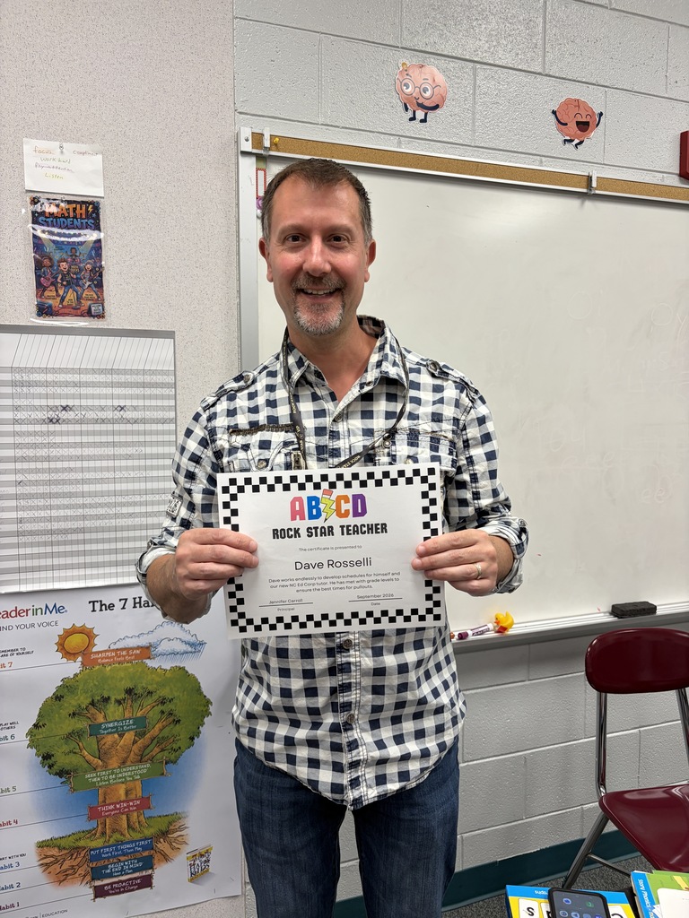 Man in a checkered shirt holding a certificate titled ABC'D. Behind him is a whiteboard and a red chair.