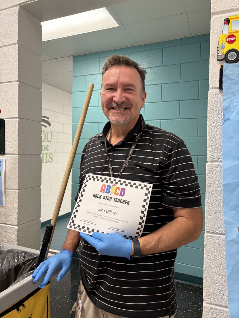 A man in a black and white shirt holds a certificate and smiles in a school hallway.