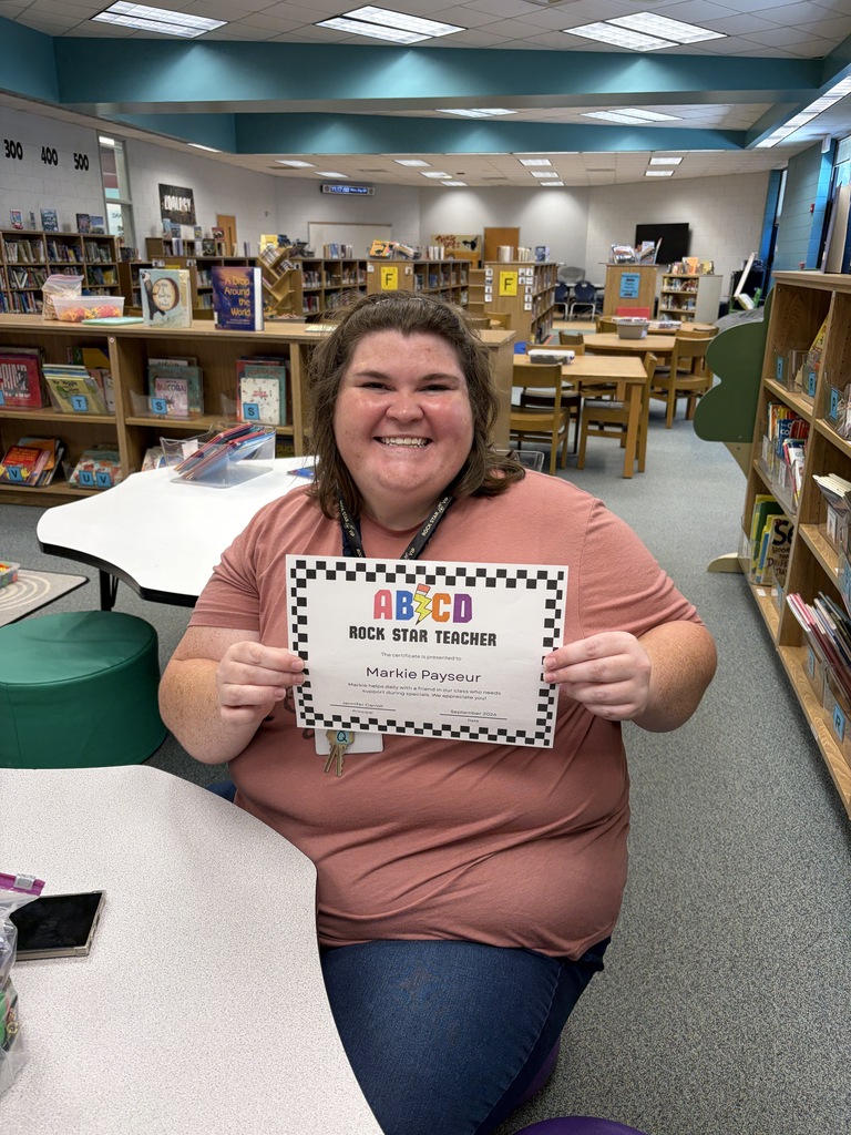 A smiling woman holds a certificate while seated at a desk in a room full of bookshelves.